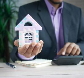 Businessman in suit holds model house, calculates real estate investments.